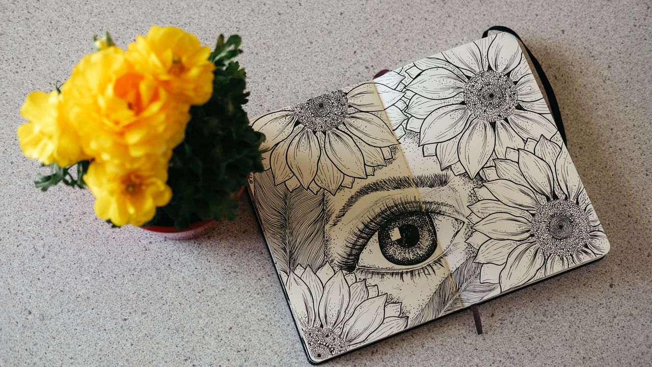 Detailed sketch of sunflowers and eye in notebook beside vibrant yellow flowers on textured surface.