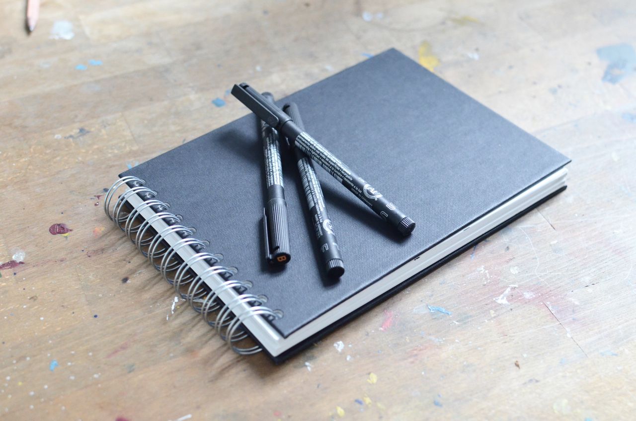 Liners and pencils placed on sketchbook with spiral on table stained with multicolored paint